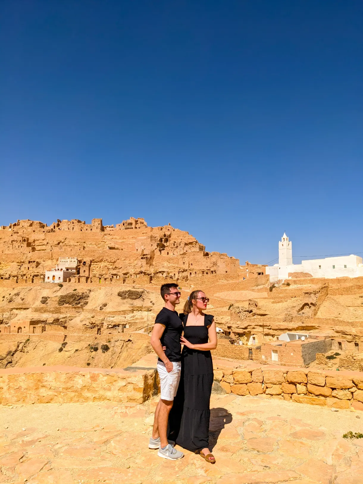Marie et Romain habillés en noir devant un village troglodyte typique tunisien Chenini avec un ciel bleu