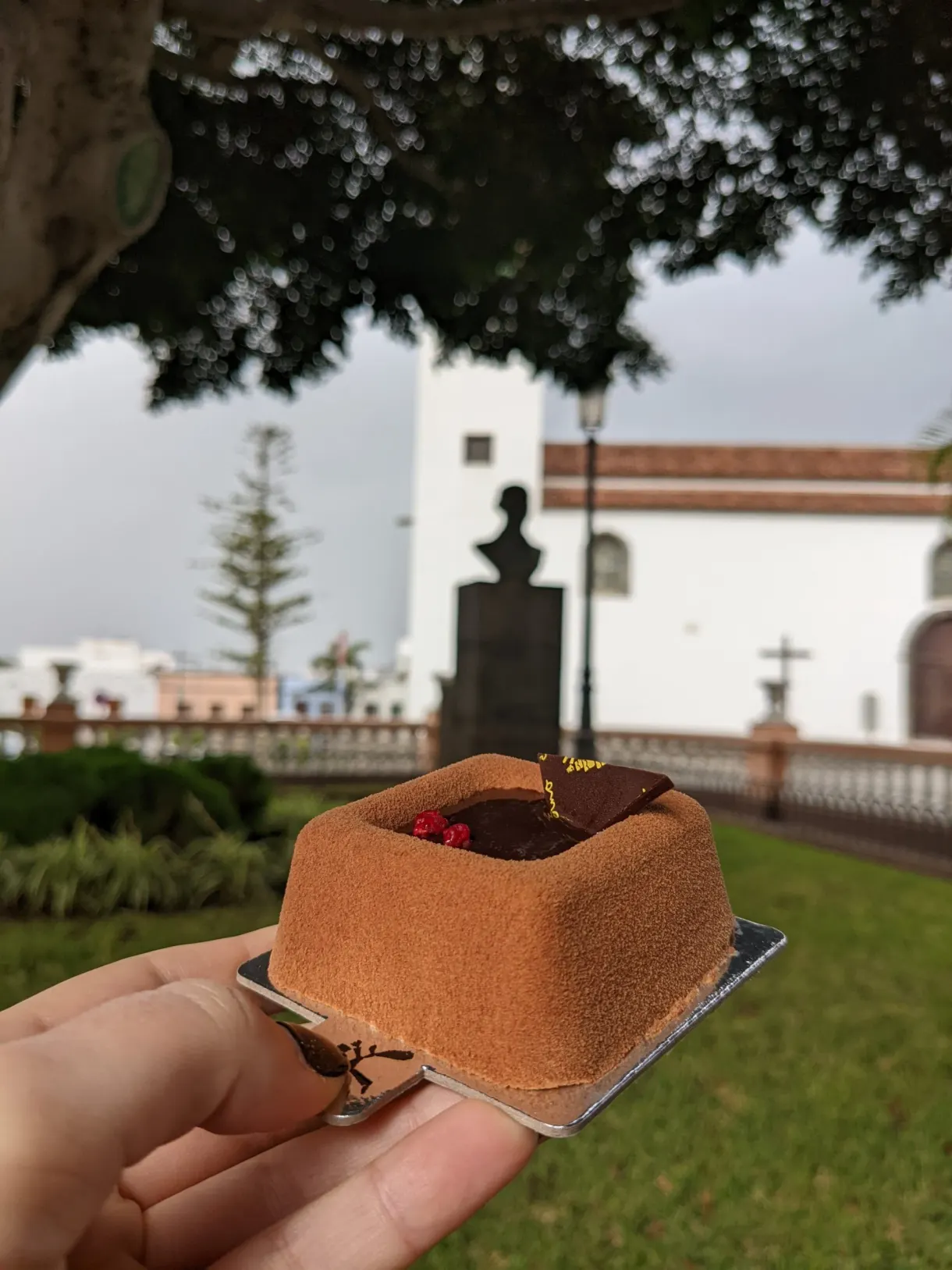 Petit gâteau au chocolat tenu devant un jardin et une église blanche