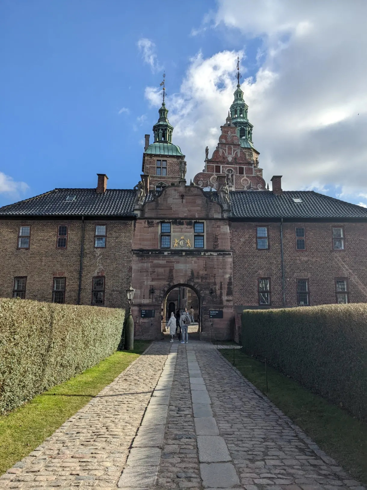 Allée pavée bordée de hautes haies menant au porche du château
