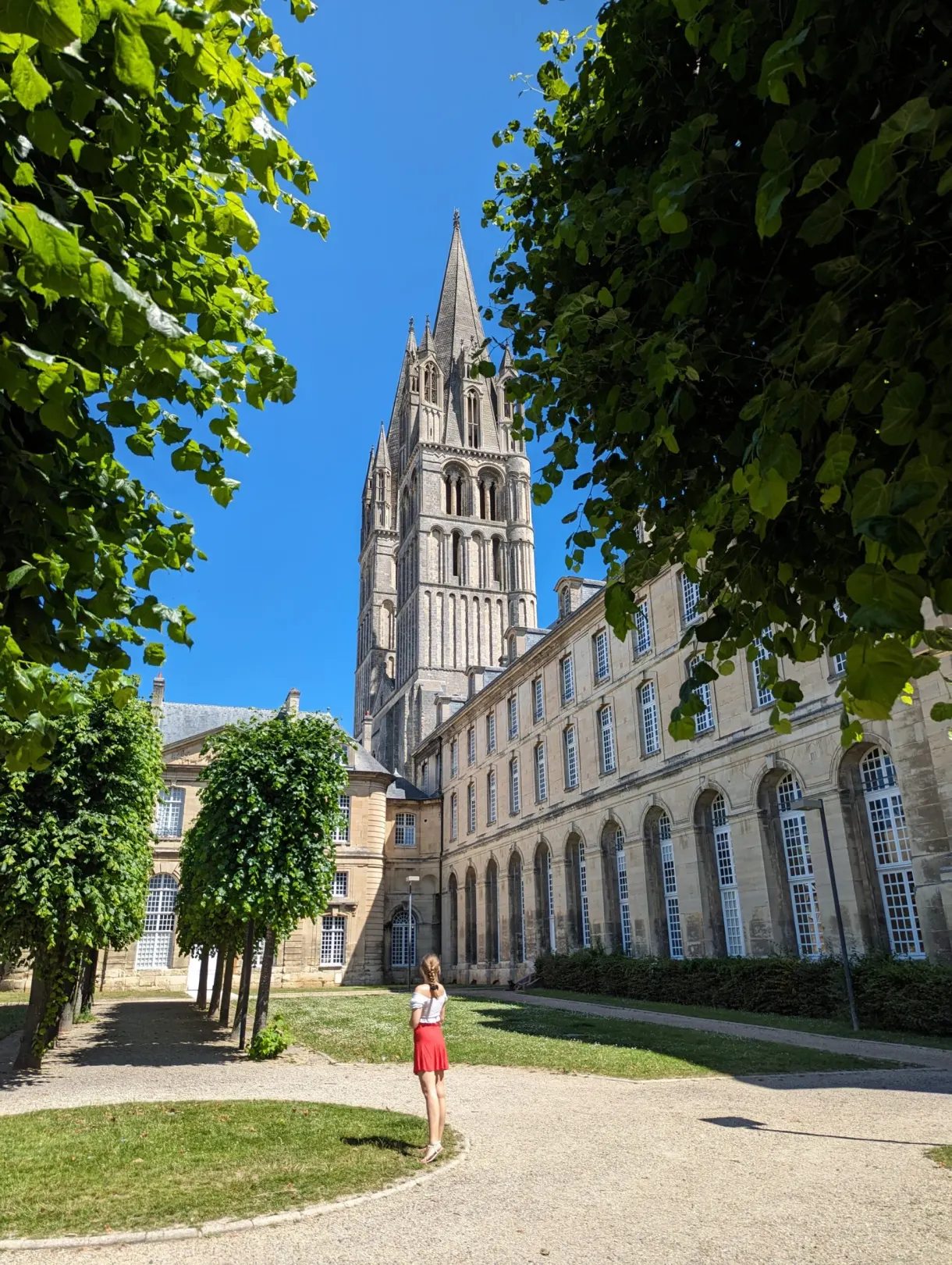 Allée d'arbres taillés longeant l'abbaye, promeneuse en robe rouge