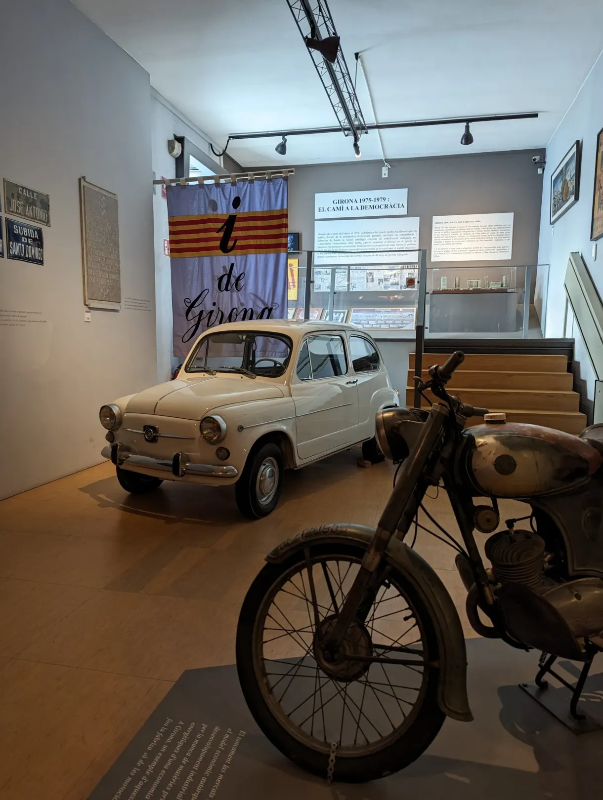 Petite voiture blanche et moto anciennes devant un drapeau catalan
